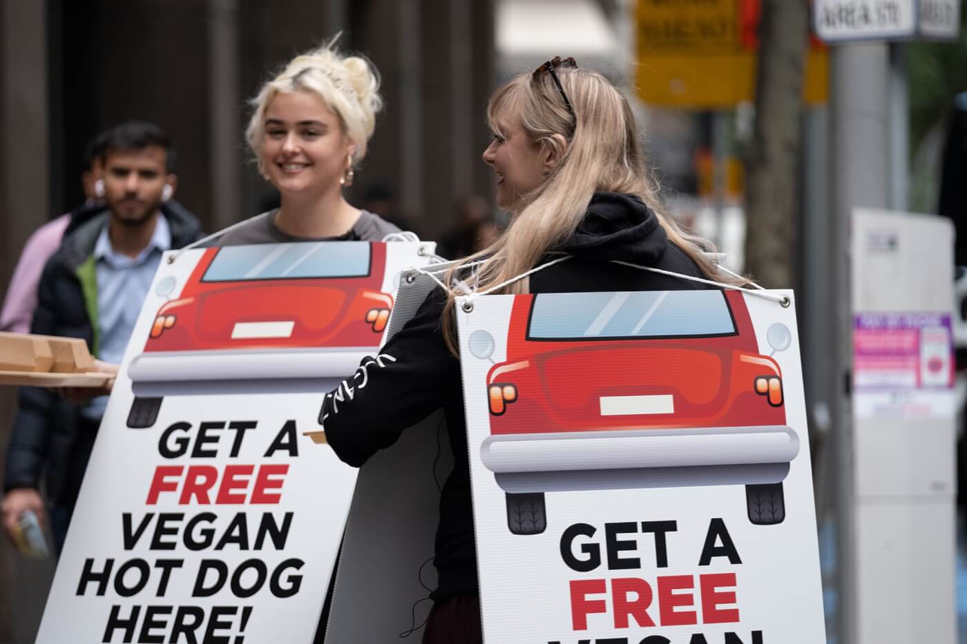 Stefania and Stace wear signs reading: Free Hot Dogs from Cars and Get a Free Vegan Hot Dog Here