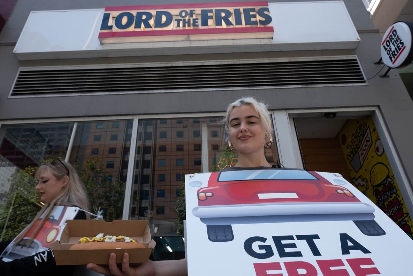 PETA activists wearing sandwich boards hand out hot dog to person on the street with "Lord of the Fries" sign in background