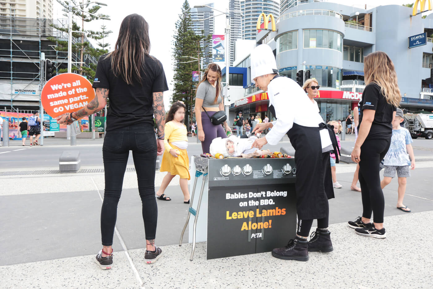 a child and adult walk past a PETA protest with a doll on a bbq. Sign reads: "babies don't belong on the BBQ. Leave lambs alone."