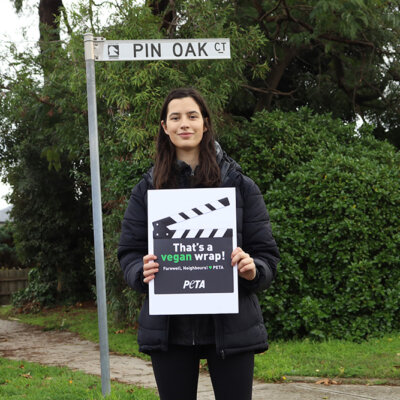 A woman stands in front of a street sign for "Pin Oak Court" the real life Ramsay Street.