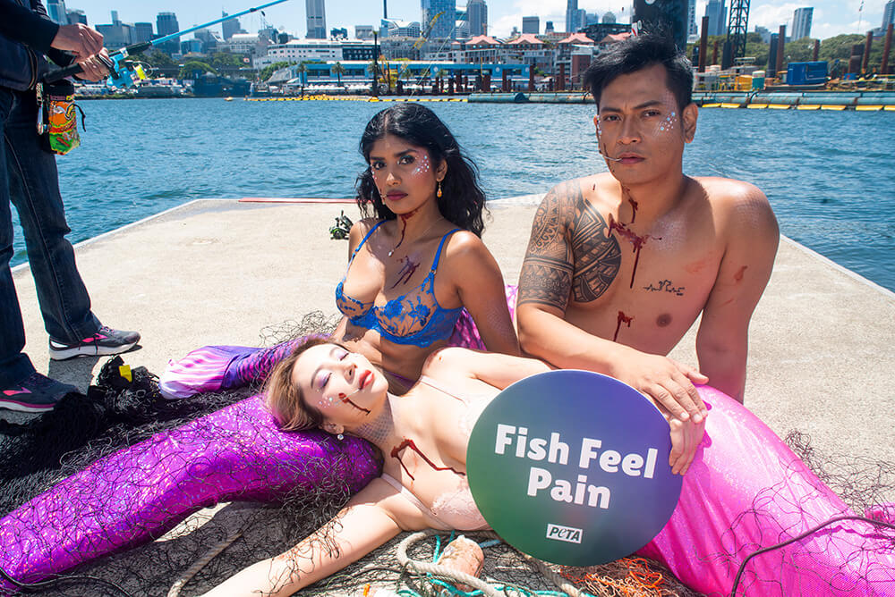 activists wearing mermaid tails and wrapped in fishing nets with hooks in their mouths.